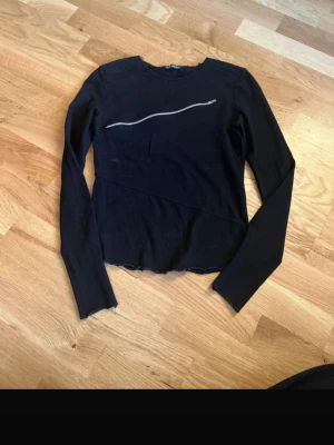 Svart långärmad topp med dragkedja - Cool svart långärmad topp från Riki med en unik, vågig dragkedja över bröstet. Toppen har en enkel, tight passform och är perfekt för dig som gillar edgy detaljer. Materialet känns mjukt och stretchigt, vilket gör den bekväm att bära.
