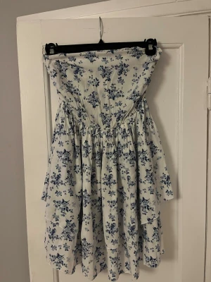 Vit blommig axelbandslös klänning - Söt axelbandslös klänning i vitt med blått blommönster. Klänningen har smock upptill för en bekväm passform och ett luftigt, volangigt kjolparti. Perfekt för varma dagar och sommarens alla tillfällen.
