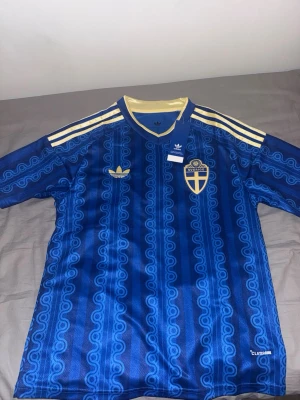 Blå Sverige fotbollströja Adidas - Säljer en blå fotbollströja från Adidas med Sveriges landslagsemblem på bröstet. Tröjan har gul krage, gula ränder på axlarna och ett mönster i olika blå nyanser. Tillverkad i lätt och ventilerande material, perfekt för träning eller match.