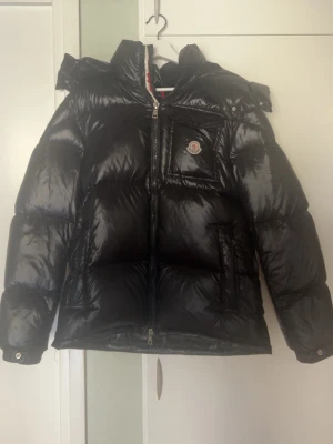Svart dunjacka från Moncler - Snygg svart dunjacka från Moncler med glansig finish, stor huva och dragkedja framtill. Jackan har en bröstficka med lock och Moncler-logga, samt två sidofickor. Perfekt för kalla vinterdagar och riktigt trendig look.