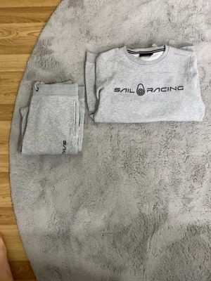 Grått loungewear-set Sail Racing (150) - Mysigt grått loungewear-set från Sail Racing med sweatshirt och matchande byxor. Tröjan har rund halsringning och svart logga framtill, byxorna har dragsko i midjan och diskret logga på benet. Perfekt för chill dagar hemma.