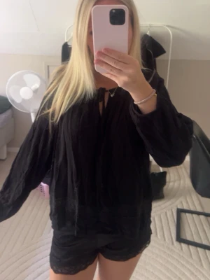 Svart blus med spetsdetaljer - Säljer en svart blus med långa ärmar och knyt vid halsen. Blusen har fina spetsdetaljer längst ner och är tillverkad i ett lätt, luftigt material. Blusen är lite söndrig längst ut på ärmarna.