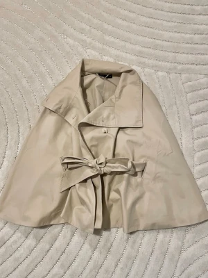 Beige cape trenchcoat med knytskärp - Hej! Säljer nu denna fina poncho kappa då den aldrig kommit till användning. OBS! (Prislappen är ej kvar) Kappan är i nyskick och i storlek XS 🤎