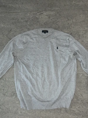 Ljusgrå tröja från Polo Ralph Lauren - Snygg ljusgrå stickad tröja från Polo Ralph Lauren med klassisk blå logga broderad på bröstet. Tröjan har rund halsringning, långa ärmar och ribbade muddar vid ärmslut och nederkant. Perfekt för en clean och stilren look.