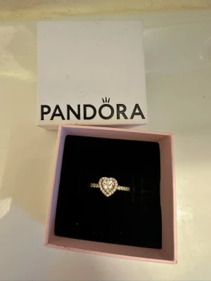 Pandora ring med hjärtformad sten - Ring med hjärta från Pandora. Webbpris: 999kr. I princip aldrig använd. Finns inget tecken på användning alls. 14 karats guldplätering. Storleken från pandoras hemsida är 54 och ringen är 17.2 mm i diameter och 54 i omkrets. Man kan även se den siffran på ringen som visas på en bild!