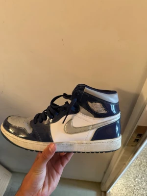 Blåvita Air Jordan 1 high  - Snygga Air Jordan 1 Mid sneakers i blått och vitt med silvriga detaljer och klassisk vinge-logga på sidan. Skorna har en hög siluett, vita paneler och blå lackdetaljer. Perfekta för dig som gillar streetwear och vill sticka ut med en ikonisk modell.