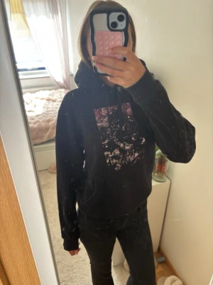 Svart hoodie med rosa tryck - Säljer en svart hoodie med huva och ett rosa, lite slitet tryck på bröstet. Hoodien har en klassisk passform med känguruficka framtill och dragsko i huvan. Perfekt för dig som gillar en avslappnad och cool stil.