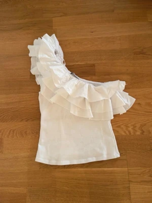 Vit oneshoulder topp med volanger - Säljer en vit oneshoulder topp med stora volangdetaljer över axeln. Toppen är ärmlös och har en snygg, asymmetrisk design som verkligen sticker ut. Perfekt för dig som vill ha något unikt och trendigt i garderoben.