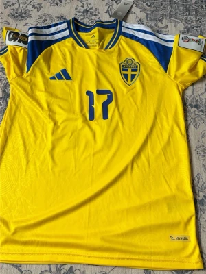Svensk landslagströja nr 17 Adidas - Gul svensk landslagströja från Adidas med blå detaljer, nummer 17 och namnet Gyökeres på ryggen. Tröjan har blåa ränder på axlarna, broderad landslagssköld och Climacool-material för bra ventilation. Perfekt för fotbollsfans!