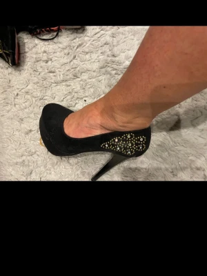 Svarta pumps med gulddetaljer - Snygga svarta pumps med hög klack och rund tå. Skorna har dekorativa guldfärgade nitar på sidan som ger en extra touch. Materialet ser ut att vara mockaimitation och klacken är smal och elegant. Perfekta för att lyfta din outfit.