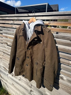 Overshirt - Snygg brun rock med dubbelknäppning, axelklaffar och dekorativa knappar vid ärmslut. Rocken har en klassisk krage och är långärmad. Tillverkad i ett robust bomullsmaterial, perfekt för höst och vår.