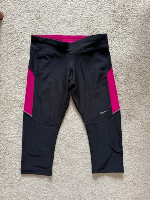 Svarta Nike Dri-FIT träningstights  - Snygga svarta träningstights i knälängd från Nike med rosa paneler på sidorna och liten reflexdetalj. Tillverkade i Dri-FIT material som andas och håller dig torr under träningen. Har en liten ficka med dragkedja bak i midjan. Perfekta för löpning eller gym. Kolla gärna mina andra annonser.