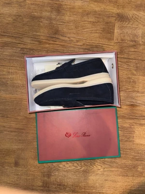 Mörkblå mocka loafers Love Roma - Snygga mörkblå loafers i mocka från Love Roma. Skorna har en klassisk siluett med dekorativ metallspänne framtill och kontrasterande vit sula. Perfekta för dig som gillar stilrena detaljer och vill ha något bekvämt men ändå trendigt.