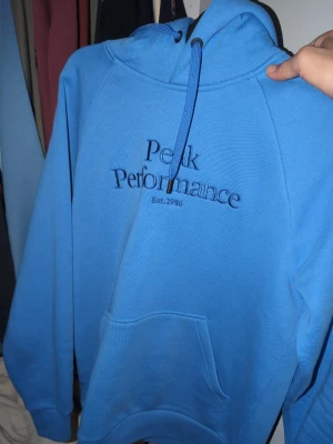 Blå hoodie från Peak Performance - Snygg blå hoodie från Peak Performance med broderad logga och text på bröstet. Klassisk känguruficka framtill, huva med dragsko och ribbade muddar. Perfekt för en avslappnad och sportig stil. .köptes förra  månaden 