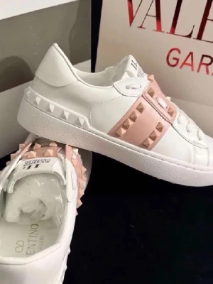 Vita sneakers med rosa nitar - Snygga vita sneakers från Valentino med breda rosa band över vristen dekorerade med ikoniska pyramidformade nitar. Skorna har en chunky sula och detaljerade sömmar för en exklusiv look. Perfekta för dig som vill sticka ut med stilrena men edgy detaljer.