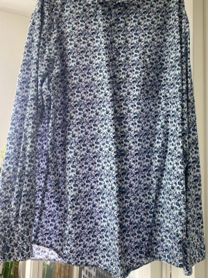 Blommig skjorta från Peter Adams XL/L - Snygg långärmad skjorta från Peter Adams i storlek XL mer som L. Skjortan har ett blommigt mönster i blått och vitt, klassisk krage och knappar framtill. Perfekt för dig som vill sticka ut lite extra med en unik och fräsch look.