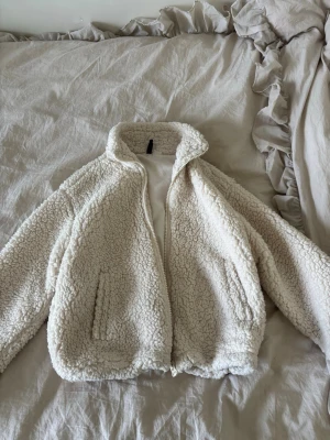 Krämvit teddyjacka med dragkedja - Mysig krämvit teddyjacka med hög krage och dragkedja framtill. Jackan har två sidofickor och är gjord i fluffigt, mjukt teddy-material som håller dig varm. Perfekt för kyliga dagar och ger en trendig look.