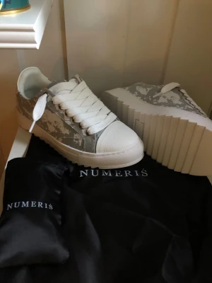 ÖPPEN FÖR BUD Vita och grå sneakers från Numeris - Snygga sneakers från Numeris i vitt och grått med coolt kamouflagemönster på sidorna. Skorna har tjock, räfflad platåsula och breda vita skosnören. Ovandelen är i slätt material och tån är rundad för en modern look.