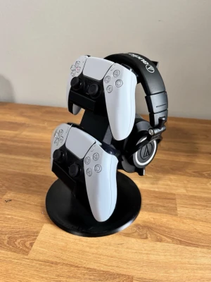 Dubbelställ för PS5-handkontroller - Snyggt svart ställ för två vita PlayStation 5 DualSense-handkontroller. Stället är gjort i plast och har plats för både handkontroller och hörlurar. Perfekt för att hålla gaminghörnan organiserad och stilren.