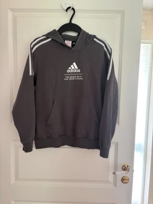 Mörkgrå hoodie från Adidas - Snygg mörkgrå hoodie från Adidas med vit logga och text på bröstet samt klassiska vita ränder på axlarna. Tröjan har huva och en stor magficka framtill. Perfekt för en avslappnad och sportig stil. Kolla gärna mina andra annonser❗️
