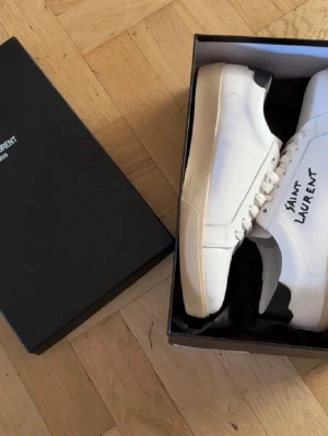 Vita Saint Laurent sneakers - Säljer ett par vita Saint Laurent sneakers i läder med svart detalj på hälen och handskriven logga på sidan. Klassisk låg modell med vita snören och beige sula. Perfekta för dig som vill ha en clean och stilren look.