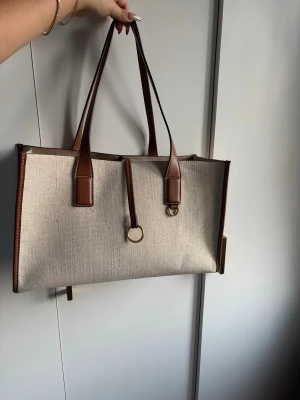 Beige/brun handväska med bruna detaljer - Stilren handväska i beige canvas med bruna läderhandtag och kanter. Väskan har rektangulär form, dekorativa guldringar framtill och en praktisk innerpåse med dragsko. Från Zara. Helt ny. Inga defekter