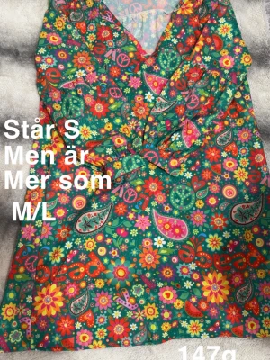 Färgglad blommig kortklänning - Kort klänning med färgglatt blommigt och paisleymönster i grönt, rosa, orange och gult. V-ringad omlottmodell med volangdetaljer och knyt i midjan. Perfekt för sommarens alla tillfällen.🌸 Sparsamt använd. Står storlek s men upplever som stor. Mer som M/L