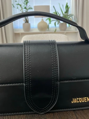 Jacquemus Le Bambino - Säljer denna stilrena Jacquemus Le Bambino  i svart med guldfärgad logotyp. En elegant och tidlös modell med handtag upptill och den karakteristiska remdetaljen framtill.  Väskan är i fint skick med mindre spår av användning, se bild för skick. Passar både till vardags och till finare tillfällen. Tillverkad av slätt läder, mått: 23,5 cm x 13 cm.