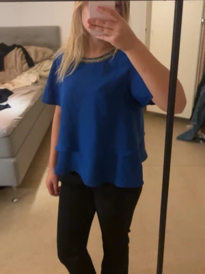 Blå topp med volang och pärldetalj - Snygg blå topp med korta ärmar och volang nedtill. Rund halsringning dekorerad med pärldetaljer som ger en extra touch. Perfekt att styla med jeans eller kjol för en trendig look.