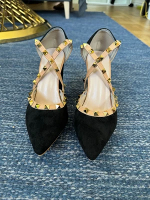 Svarta pumps med nitar och remmar - Snygga svarta pumps med spetsig tå och beige remmar dekorerade med guldfärgade nitar. Skorna har en elegant klack och korsade remmar över foten som ger en cool och trendig look. Perfekta för dig som vill sticka ut med din stil.