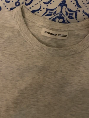 Ljusgrå t-shirt från Pull&Bear - En basic ljusgrå t-shirt från Pull&Bear med rund hals och korta ärmar. Tillverkad i mjuk bomull för en skön och avslappnad känsla. Perfekt att matcha med jeans eller shorts för en enkel och clean look.
