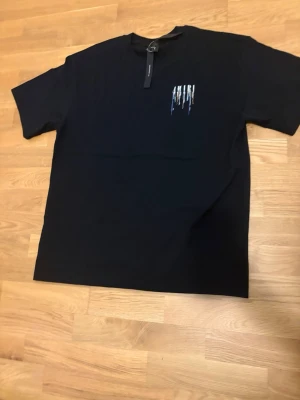 Svart AMIRI t-shirt med färgstänk - Säljer en svart t-shirt från AMIRI med färgstänk och logga på bröstet samt stor färgglad AMIRI-text med drip-effekt på ryggen. T-shirten har korta ärmar och är gjord i mjuk bomull. Perfekt för dig som gillar streetwear och vill sticka ut.