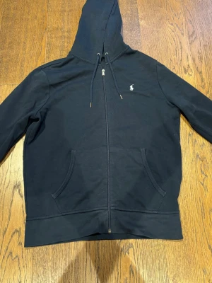 Svart hoodie med dragkedja Polo Ralph Lauren - Klassisk svart hoodie från Polo Ralph Lauren med dragkedja framtill, två fickor och justerbar huva med snören. Liten vit logga broderad på bröstet. Tillverkad i mjukt bomullsmaterial som är skönt att bära.
