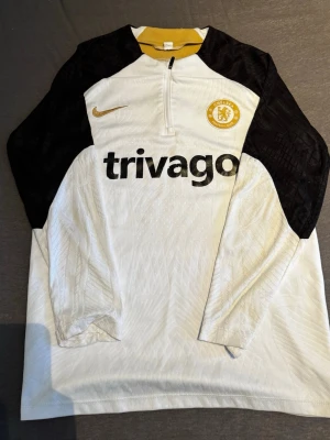 Chelsea FC vit träningströja Nike - Chelsea FC långärmad träningströja från Nike i vitt med svarta ärmar och gul detalj vid kragen. Halv dragkedja framtill, klubbmärke och Nike-logga på bröstet samt 'trivago' tryckt över bröstet. Tillverkad i lätt och ventilerande material.