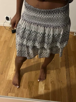 Lagom kort kjol med zigzag-mönster! - Supersnygg kort kjol med svartvitt zigzag-mönster och bred smock i midjan. Kjolen har en luftig och flowy passform som ger en avslappnad vibe. Perfekt till sommaren och lätt att matcha med olika toppar.