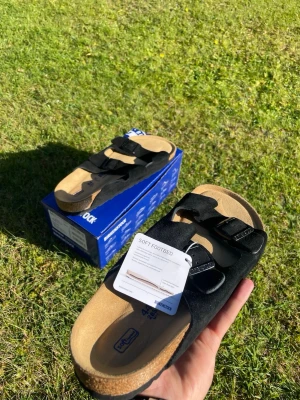 Svarta Birkenstock sandaler med spännen - Säljer ett par svarta Birkenstock sandaler med två justerbara spännen och mjuk fotbädd. Ovandelen är i mocka och sulan är i kork och gummi för extra komfort. Perfekta för dig som vill ha både stil och bekvämlighet. De är riktigt populära och vid snabb affär sänker jag priset, säljs billigt av en anledning 