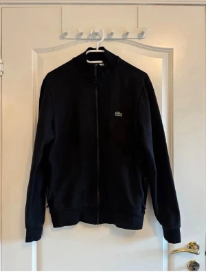 Svart zip hoodie från Lacoste - Snygg svart hoodie från Lacoste med dragkedja framtill och klassisk krokodil-logga på bröstet. Tröjan har hög krage, långa ärmar och ribbade muddar. Tillverkad i mjukt material som passar perfekt till en avslappnad stil.