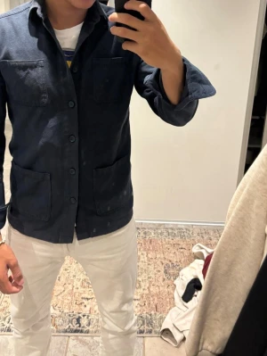 Mörkblå overshirt - Stilren mörkblå overshirt med fyra stora fickor framtill och knäppning hela vägen. Overshirten har klassisk krage och raka ärmar. Perfekt att bära över en t-shirt för en avslappnad men snygg look. Materialet ser ut att vara mjukt och bekvämt.