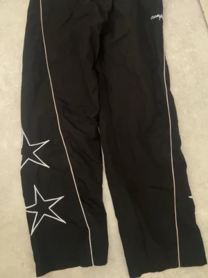 Carpenter byxa med stjärnor - Svarta byxor från Carpenter med vita stjärnor broderade på benen och vita piping-detaljer längs sidorna. Byxorna har resår i midjan och är i en avslappnad passform. Perfekta som streetwear eller till vardags! Säljes i storlek M.