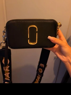 Svart axelväska Marc Jacobs - Snygg svart axelväska från Marc Jacobs med guldigt logodetalj framtill. Väskan har en bred axelrem med text i guld och vitt, samt justerbar längd. Tillverkad i ett tåligt material med dragkedja upptill. Perfekt för att bära det viktigaste med stil.