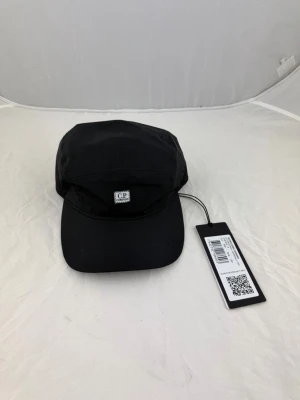 Svart keps från C.P. Company - Snygg svart keps från C.P. Company med klassisk fempanelsdesign och liten logotyp framtill. Kepsen har böjd skärm och justerbar passform. Tillverkad i ett slitstarkt material som passar perfekt till streetwear-looken. 