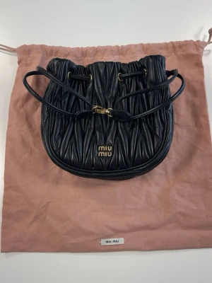 Svart handväska från Miu Miu - Säljer en svart handväska från Miu Miu i mjukt, vadderat läder med snyggt veckad design. Väskan har dragskostängning med guldfärgade detaljer och justerbar axelrem. Perfekt för dig som vill ha en trendig och lyxig accessoar.0693