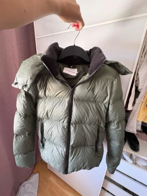 Olivgrön dunjacka med huva - Snygg olivgrön dunjacka med stor huva och dragkedja framtill. Jackan har två sidofickor med dragkedja och quiltad design som ger en puffig look. Perfekt för kalla vinterdagar och riktigt varm. Passar dig som vill ha en stilren och praktisk vinterjacka.