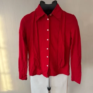 Phard Vintage Red Button-Up Blouse with Tie Collar - Passar: S  Mått (plagget liggande platt): Bröstmått (rakt över, axel till axel): 40 cm Ärmlängd: 57 cm Totallängd: 52 cm  Skjortblus från italienska märket Phard i klarröd färg med figurnära passform, spetsig krage och genomgående knappknäppning. Distinkt detalj med ett smalt, hängande halsband/skärp i samma tyg fäst vid kragen. Tunt, rörligt jerseytyg.  Mycket gott begagnat skick – inga hål, revor eller fläckar.  PRODUKTINFO: Kategori: Dam > Blusar & Skjortor Material: 100% Polyamid 0373 