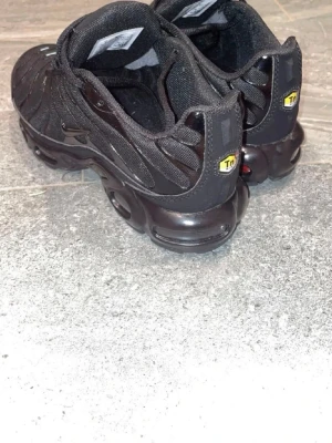 Nike Air Max Plus helsvarta sneakers - Säljer ett par helsvarta Nike Air Max Plus sneakers med TN-detaljer på hälen och klassisk vågig design. Skorna har mesh- och syntetmaterial, synlig Air-dämpning i sulan och snörning framtill. Perfekta för dig som gillar streetwear och sportig stil.