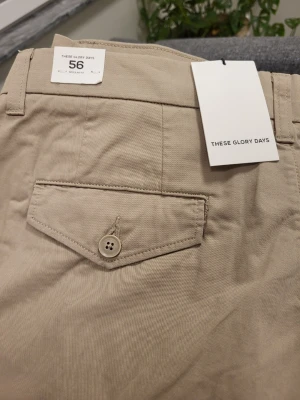 Beige cargobyxor These Glory Days - Snygga beige cargobyxor från These Glory Days med raka ben och flera praktiska fickor, inklusive sidofickor med lock och knapp. Tillverkade i ett mjukt bomullsmaterial som ger en avslappnad och modern look. Perfekta för dig som gillar stilrena och funktionella plagg.