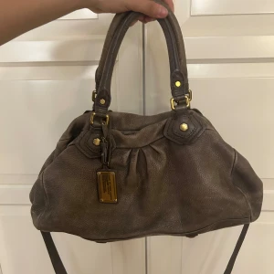 Marc Jacobs väska  - Brun/beige Marc Jacobs handväska med gulddetaljer! Väskan är i den större modellen och rymmer dator och liknande! Mycket bra skick och inga slitningar. Längre band medföljer!