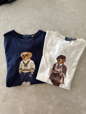 Polo Ralph Lauren t-shirts med björn - Två snygga t-shirts från Polo Ralph Lauren i bomull, en marinblå och en vit, båda med ikoniska björntryck framtill. Den marinblå har en sportig björn med solglasögon och den vita har en björn i klassisk outfit. Perfekt för dig som gillar streetstyle och preppy vibes.