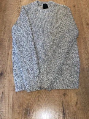 Gråmelerad stickad tröja - En stilren gråmelerad stickad tröja med rund hals och långa ärmar. Tröjan har en klassisk ribbstickad struktur och är perfekt för kyligare dagar. Passar till både jeans och kjol för en avslappnad look.