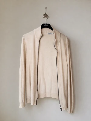 Cardigan - Ljusbeige ziptröja med ståkrage och långa ärmar. Tröjan har en enkel och stilren design med ribbade muddar vid ärmslut och nederkant. Perfekt för lager på lager och passar till det mesta.